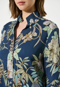Blusa blu navy con motivo floreale in colori tenui. Tessuto morbido, colletto rialzato e dettaglio dei bottoni sulla parte anteriore.