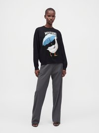 Γυναίκα που φοράει μαύρο πουλόβερ Moschino με γραφικό μιας άσπρης χήνας που κρατά μια μπλε ομπρέλα, σε συνδυασμό με φαρδιά γκρι παντελόνια και μαύρα σανδάλια.
