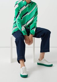 Personne assise sur un tabouret blanc portant une chemise à rayures vertes, blanches et noires, un jean bleu foncé et des baskets blanches avec des semelles vertes.