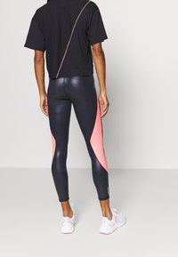 T-shirt nera cropped con una cucitura diagonale rosa, abbinata a leggings neri lucidi a vita alta con pannelli laterali rosa. Sneakers bianche.