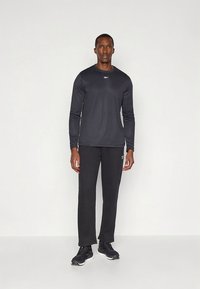 Svart langermet skjorte og svarte bukser laget av lett materiale. Skjorten har en logo; buksene har et subtilt design. Brukes med svarte joggesko.