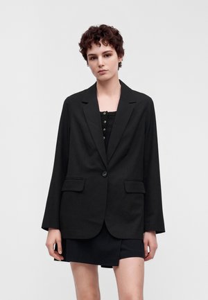 Ung person med kort krøllet hår iført en sort oversized blazer og en sort knaplukket top mod en ensfarvet lys baggrund.