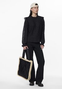Svart teksturert genser med volanger på ermene, kombinert med svarte bukser og en kviltet svart tote-bag med en kremfarget kant.