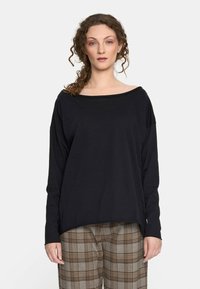 Kobieta z kręconymi włosami, ubrana w luźny czarny top z długim rękawem i brązowe spodnie w kratę, stojąca na tle jasnej, gładkiej ściany.