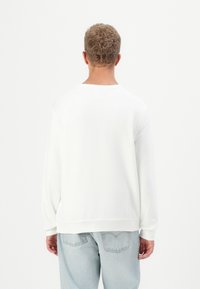Sudadera blanca de manga larga, con puños acanalados y cuello redondo, combinada con jeans de mezclilla azul claro, vista desde la parte de atrás.