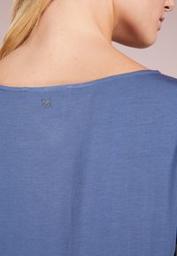 Modrá krátká rukávová blůza s širokým výstřihem. Malý motýlek z kamínků je umístěn na zadní části krku. Měkká textura.
