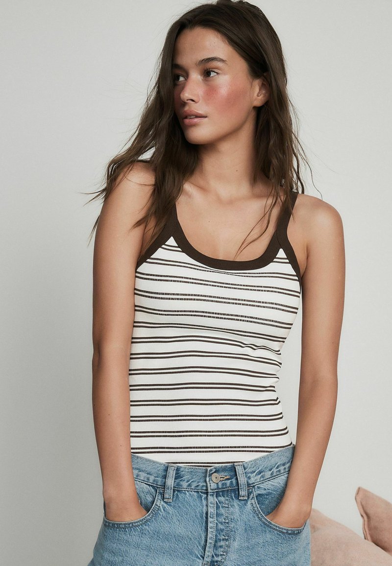 Jeune femme aux longs cheveux bruns portant un débardeur blanc rayé avec bordure marron et un jean bleu, mains dans les poches, regardant sur le côté.