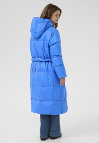 Zila pūkainā jaka ar kapuci, garām piedurknēm, elastīgu jostasvietu, quilted tekstūru un sānu šķēlumu. Kombinēta ar džinsu biksēm un melniem apaviem.
