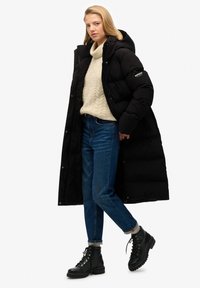 Abrigo de plumas largo y negro con capucha, usado sobre un suéter de punto de cable color crema y jeans azules. Botas negras con cordones completan el atuendo.