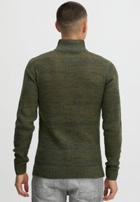 Strickpullover in Dunkelgrün mit gerippter Textur, hohem Kragen und langen Ärmeln. Hat eine figurbetonte Silhouette und dezente Farbverläufe.