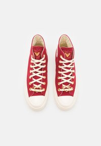 Czerwone wysokie sneakersy z płótna z białymi gumowymi podeszwami, z złotymi detalami wokół oczek i sercem na języku. Sznurówki są białe.