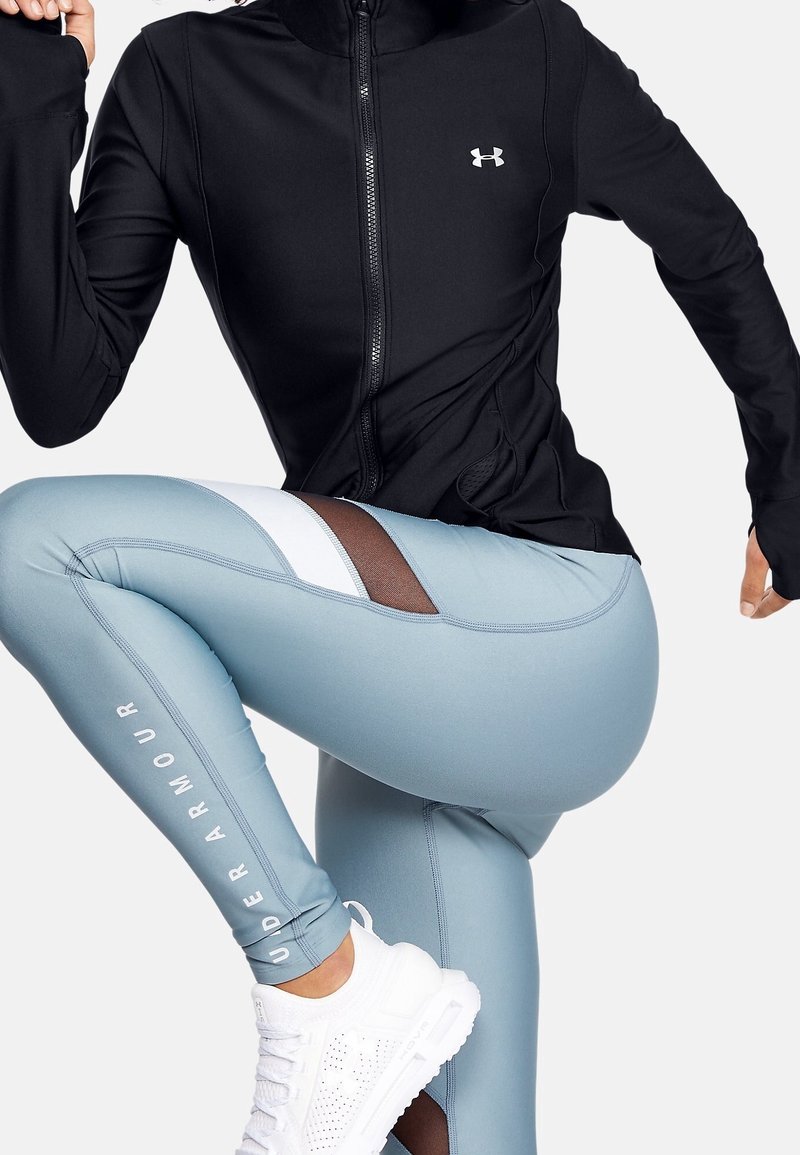 Svart figursydd jacka, blå leggings med vita och mesh-accenter, kombinerat med vita sportskor. Rena linjer och lättviktig tygsynlighet.