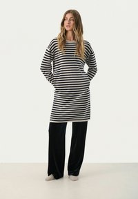Tunika a righe in crema e blu navy, con maniche lunghe e tasche laterali, abbinata a pantaloni in velluto nero e calzini chiari.