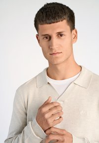 Ljusbeige stickad tröja med V-ringning och ribbade ärmslut. Bärs över en vit t-shirt och har en diskret logga på bröstet.