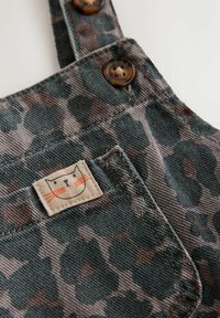 Latzhose mit Leopardenmuster aus dunkelgrünem und braunem Stoff, versehen mit Knöpfen und einem kleinen Etikett mit Katzenmotiv auf der Vorderseite der Tasche.