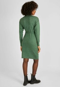 Vestito verde lavorato a maglia con vita alta, maniche lunghe e scollatura rotonda. Tessuto strutturato e lunghezza al ginocchio. Indossato con stivali neri alla caviglia.