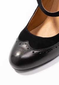 Zwarte leren en suède schoen met een ronde neus, gegolfde rand en decoratieve perforaties. Voorzien van een bandje over de enkel.