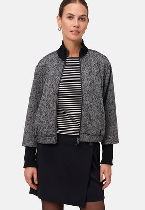 Femme portant une veste zippée à motifs sur un haut à rayures noires et blanches, associée à une jupe portefeuille noire et des boucles d'oreilles en or.