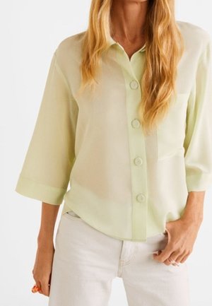 Žena v lehce žluté košili na knoflíky s tříčtvrtečními rukávy a bílých kalhotách, stojící s jednou rukou v kapse.