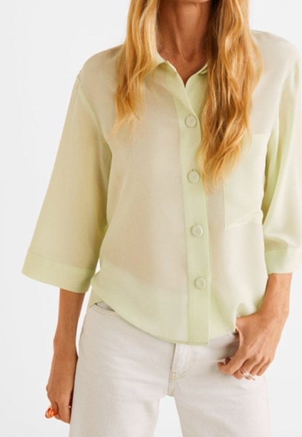 Femme portant un chemisier jaune clair à boutons avec des manches trois-quarts et un pantalon blanc, debout avec une main dans la poche.