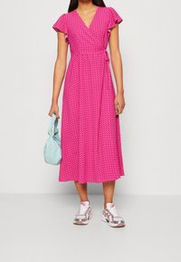 Lyserød slå-om-kjole med flagrende ærmer, geometrisk mønster og længde til midt på læggen. Kombineret med en lyseblå taske og sporty sneakers.