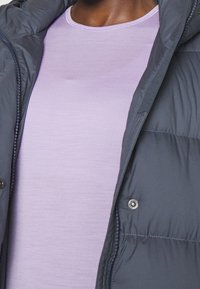Veste matelassée gris foncé avec fermeture éclair et détails en bouton, superposée à une chemise ajustée violet clair. Tissu lisse, design épuré.