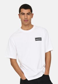 White cotton T-shirt with short sleeves, featuring a rectangular black logo patch that reads "oasis" on the left chest. Classic crew neck design.