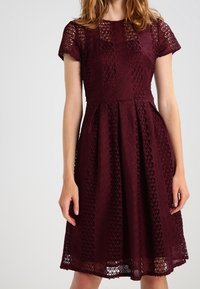 Burgunderfarbenes Spitzenkleid mit kurzen Ärmeln, tailliertem Oberteil und ausgestellt Rock, ausgestattet mit ausgeschnittenen Diamantmustern und einer strukturierten Oberfläche.