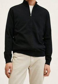 Sudadera negra con cremallera y cuello alto, puños y dobladillo acanalados, hecha de un tejido de punto suave; llevada sobre pantalones de color claro.