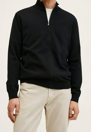 Sudadera negra con cremallera y cuello alto, puños y dobladillo acanalados, hecha de un tejido de punto suave; llevada sobre pantalones de color claro.