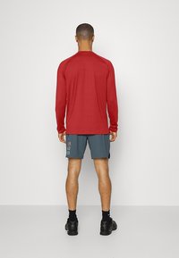 Langärmliges rotes Sportoberteil aus leichtem Material, kombiniert mit grauen Shorts mit einem bedruckten Muster. Schwarze Turnschuhe vervollständigen das Outfit.