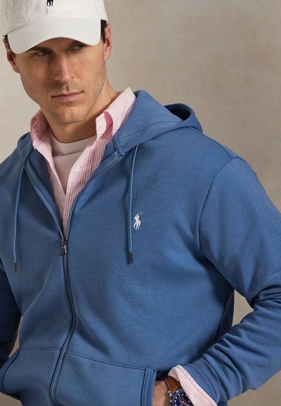 Mees, kes kannab sinist lukuga dressipluusi roosa särgi peal, valge nokkmütsi ja käekellaga, vaatab küljele ühevärvilise tausta ees.