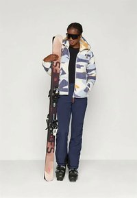 Giacca in pile con motivo geometrico blu e bianco, abbinata a pantaloni da sci neri. Il modello tiene gli sci con attacchi, indossando occhiali da sole scuri.