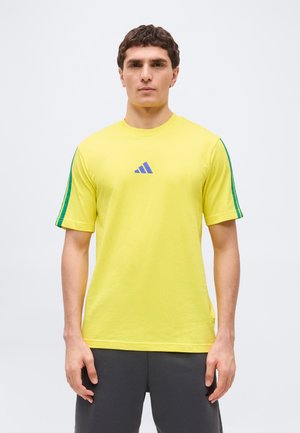 Muž v žltej krátkosleeve košeli so zelenými pruhmi na rukávoch a modrým logom, v kombinácii s tmavosivými nohavicami, stojaci na obyčajnom pozadí.