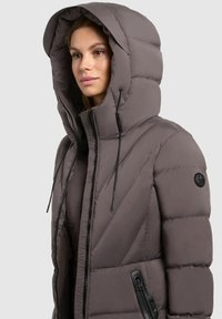 Naine kannab halli pehmendatud talvejope, millel on suur kapuuts ja musta värvi tõmbluku detailid, seistes kerge vasakule küljele heledal taustal.