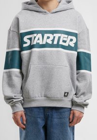 Sudadera gris con bolsillo frontal, que presenta un logo "STARTER" en color teal y rayas blancas en las mangas. Material de mezcla de algodón, ajuste relajado.