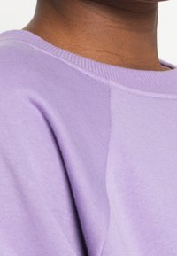 Levandulová mikina s pruhovaným límcem, která má měkkou bavlněnou texturu a volný střih. Viditelný detail švu na rameni.