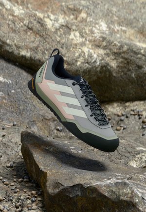 Pantof de alpinism gri cu dungi albe, șireturi negre și talpă de cauciuc. Prezintă o suprafață texturată și un logo pe călcâi. Proiectat pentru utilizare în aer liber.