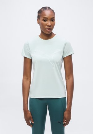 Femme aux cheveux tressés portant un t-shirt Puma bleu clair et des leggings Puma couleur sarcelle, debout devant un fond blanc uni.