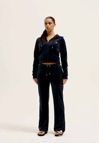 Dres z weluru w kolorze granatowym składający się z krótkiej bluzy na suwak i spodni z wysokim stanem. Z metalowymi, srebrnymi elementami i ozdobnymi sznurkami. Płaskie sandały.