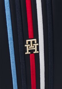 Logo en métal doré avec deux lettres H entrelacées sur un tissu sombre avec des rayures verticales bleu clair, rouge et blanc cousues.