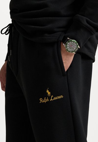 Čierne teplákové nohavice so zlatým výšivkou "Ralph Lauren" a logom, so bočnou kapsou, doplnené hodinkami s zelenými akcentmi.