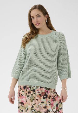 Hosszú barna hajú nő világos zöld kötött pulóverben és virágmintás midi szoknyában, rózsaszín, zöld és bézs árnyalatokkal.