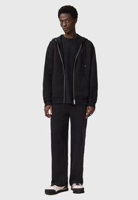 Haut de survêtement noir zippé avec poches, porté sur un t-shirt noir et un pantalon noir, présentant une coupe décontractée et une texture lisse. Baskets noires et blanches.