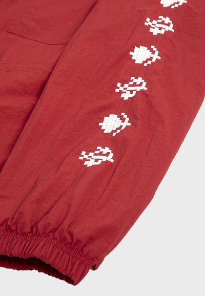 Manche en tissu rouge avec un poignet élastique et une rangée verticale de broderies florales pixélisées blanches le long du bras.