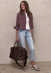 Woman in purple jacket, white shirt, light jeans, and sneakers holding a large brown duffel bag, standing against a plain wall.