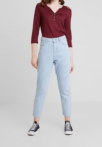 Burgundy långärmad topp med V-hals och tre knappar, kombinerad med ljusblå högmidjade jeans och svarta sneakers.