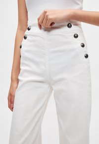 Λευκό παντελόνι με ψηλή μέση από βαμβάκι με ομαλή υφή, με μεγάλα μαύρα κουμπιά κατά μήκος των πλευρών για λεπτομέρεια.