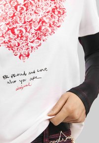 Valge T-särk, millel on südamekujuline muster punastes toonides, musta ja punasega tikitud tekst ääre lähedal ning keha ümber sobivad mustad varrukad.
