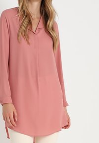 Långärmad, ljusrosa blus i ett mjukt tyg. Har krage, knäppta ärmslut och en rundad nederkant för en avslappnad passform.